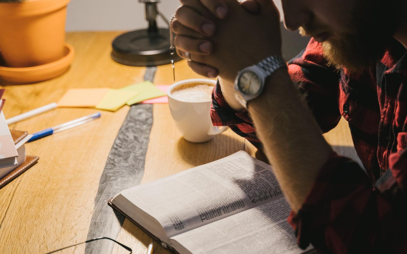 A man praying with hands clasped over an open Bible on a wooden table